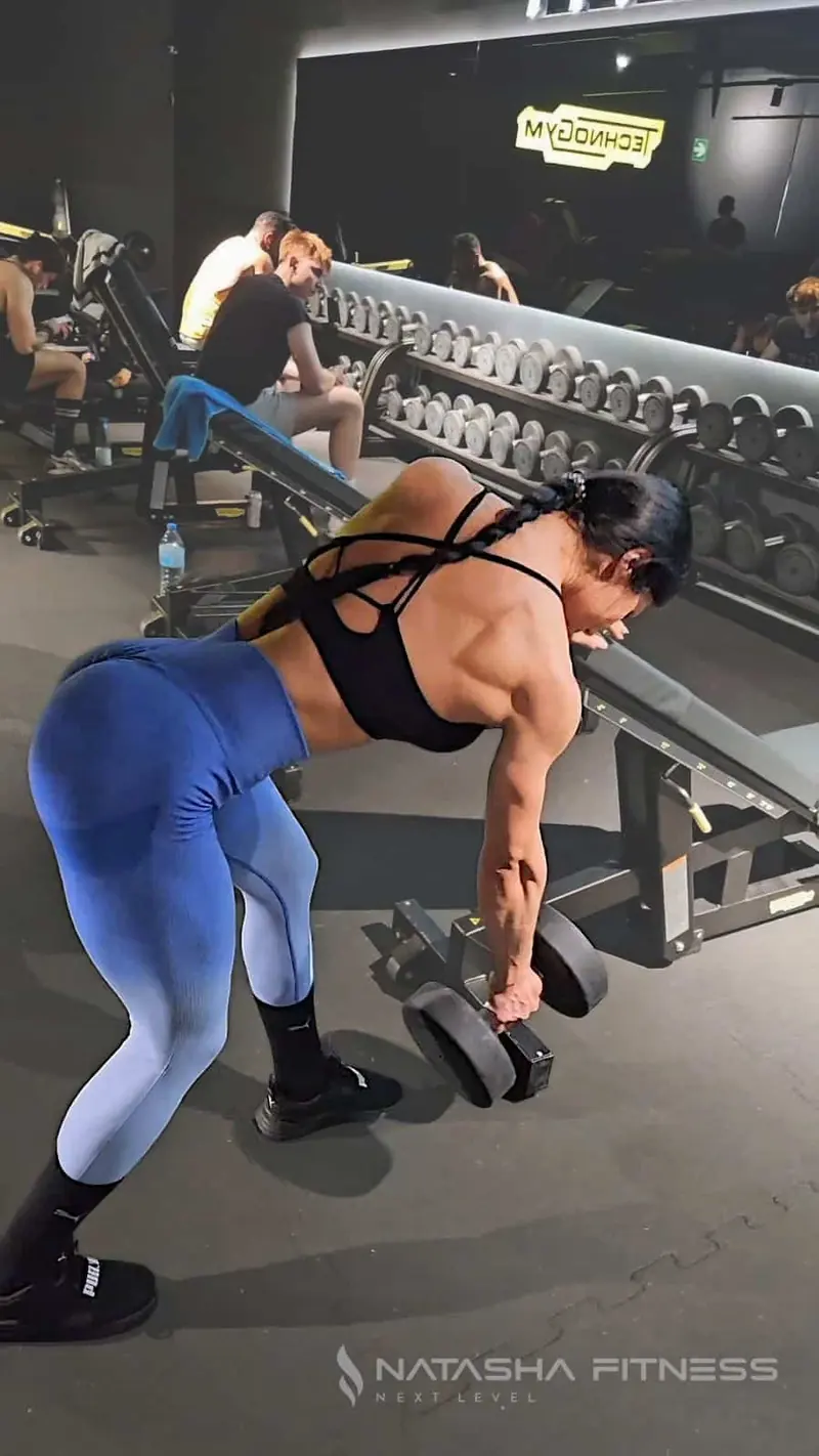 Natasha entrena inclinada hacia delante con una mancuerna en cada mano, junto a un banco y el rack de mancuernas